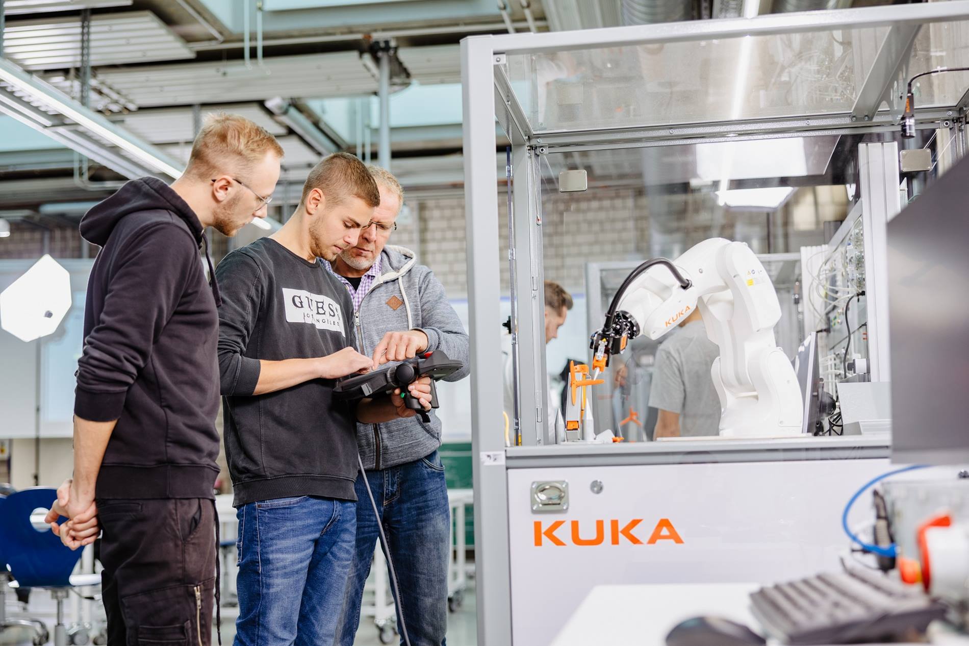Three students from the Gewerbliche Schule Göppingen standing in front of a networked KUKA robot training cell. 
