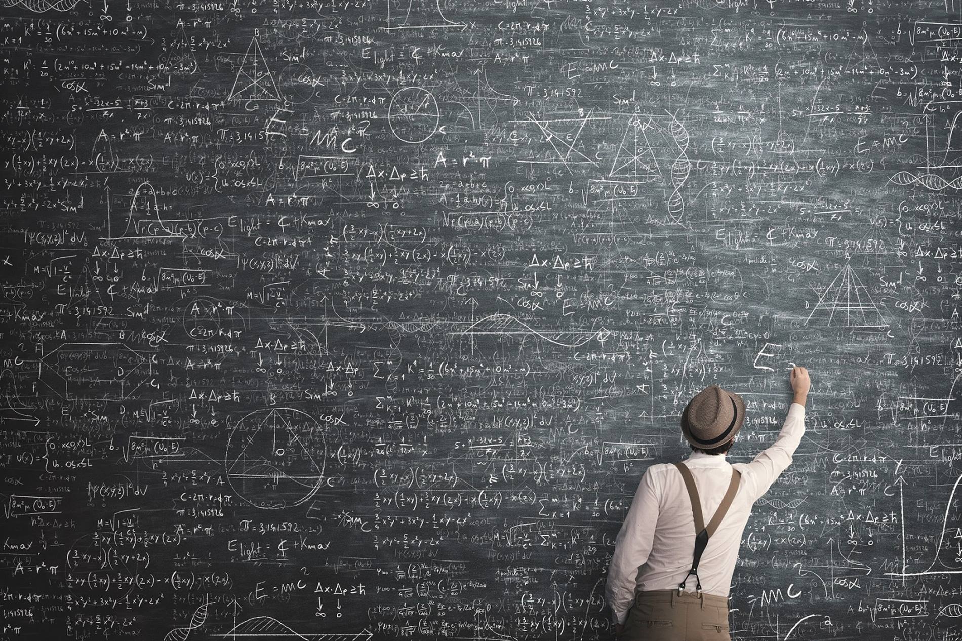 Young man stands in front of a large blackboard and writes mathematical equations with a chalk.