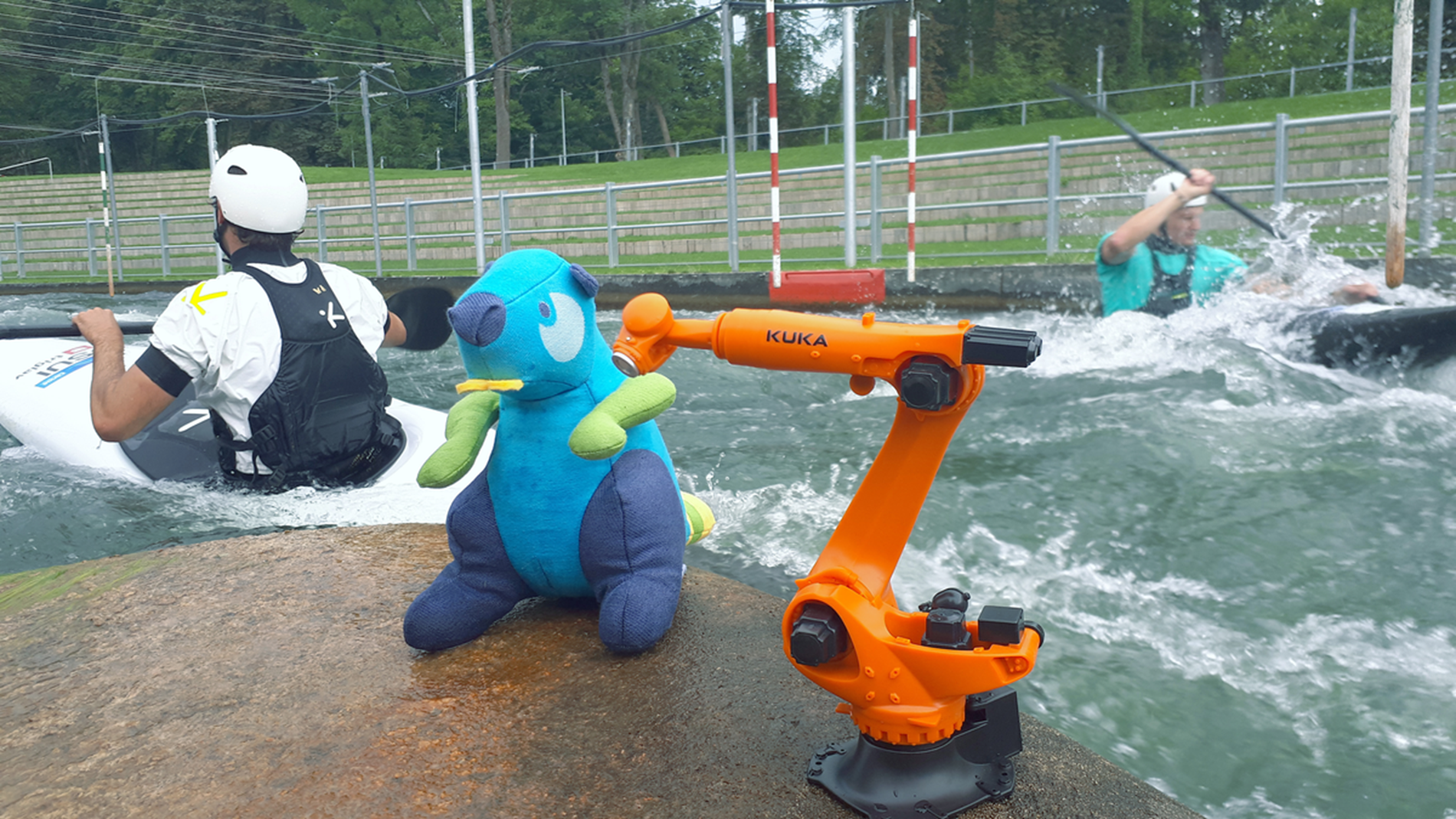 Gustl, the official mascot of the World Canoe Championships, and a KUKA robot model in front of two canoeists in Augsburg's Eiskanal.