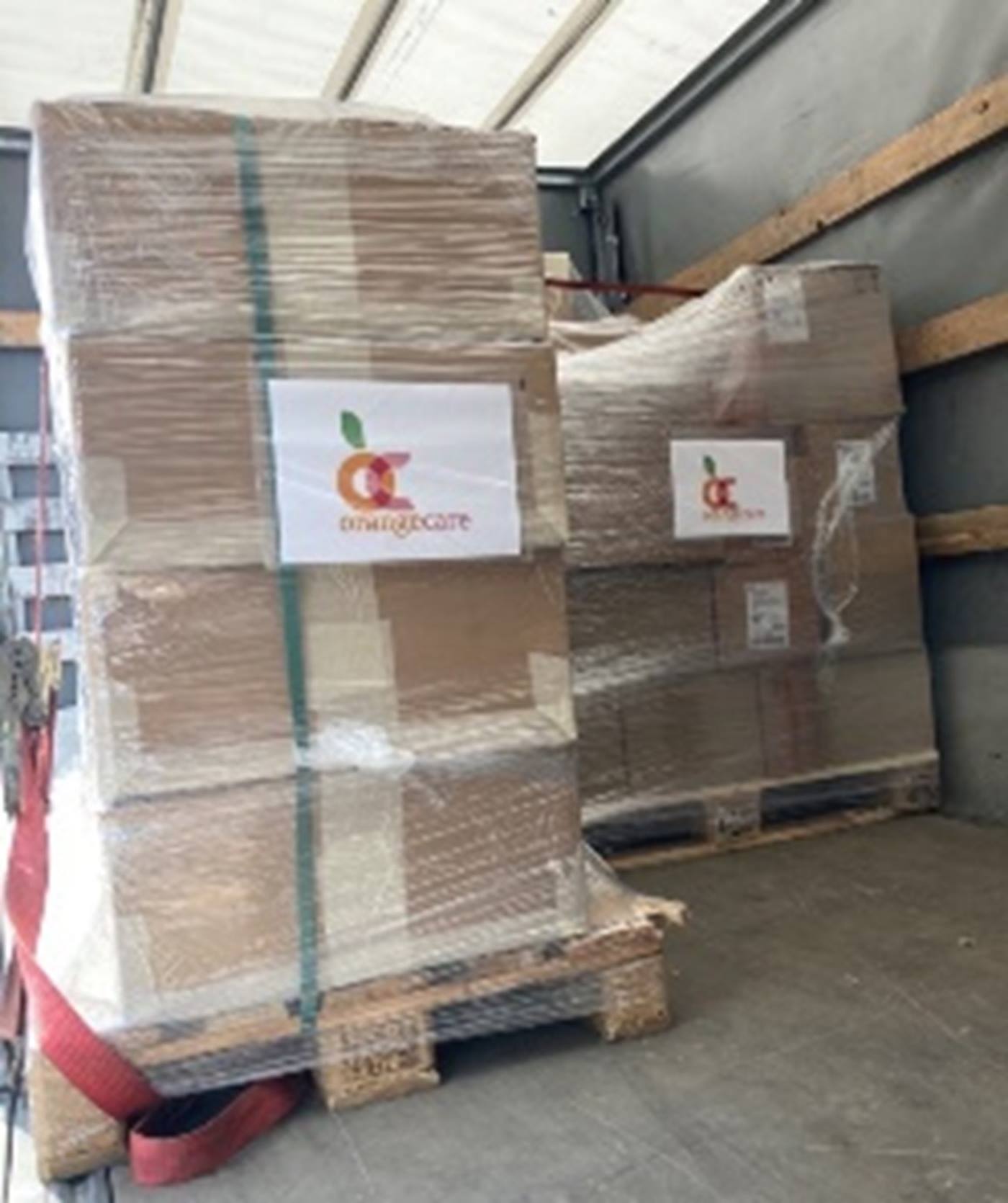 Two pallets full of cardboard boxes with relief supplies, marked with an Orange Care logo, are loaded onto a truck. 