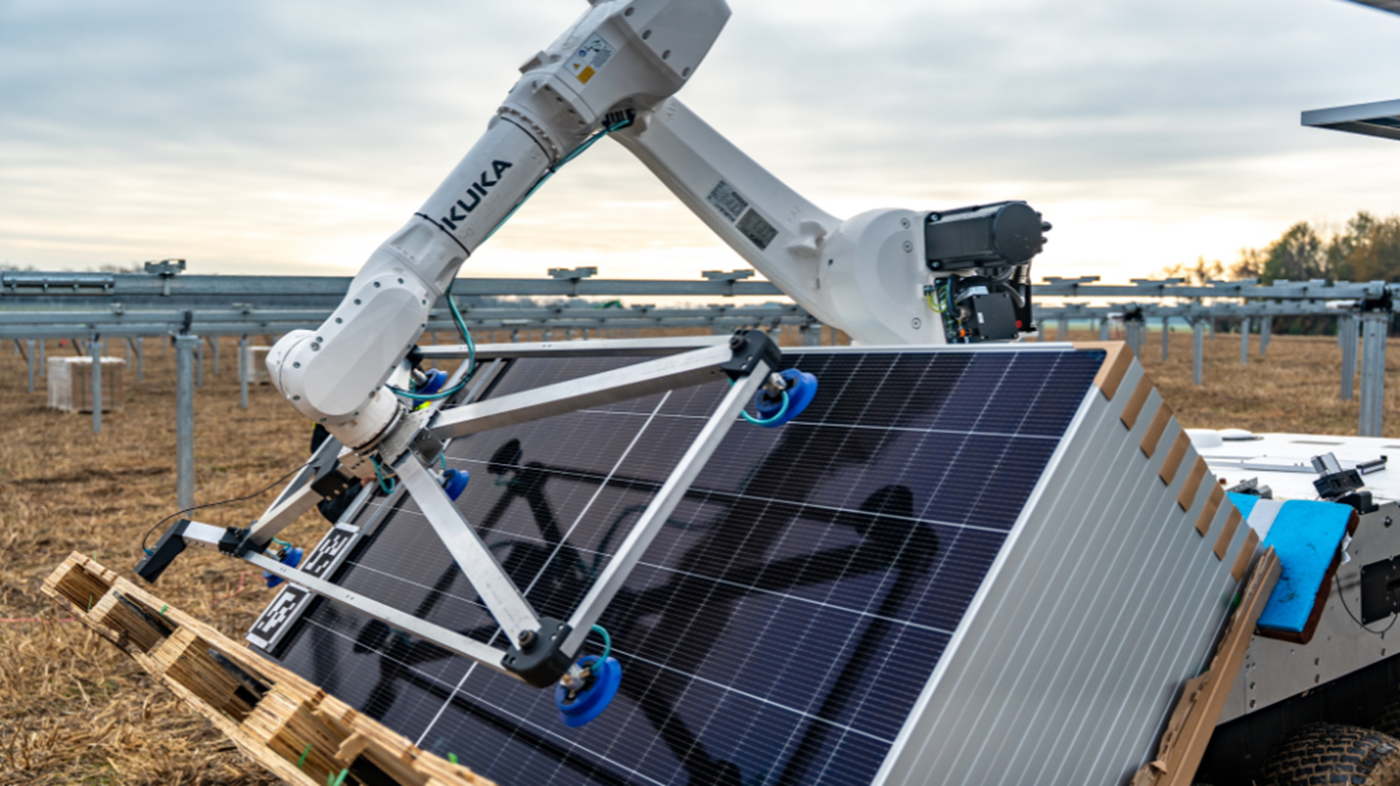 A KUKA robot lifts and aligns a solar module while installation workers secure it to a tracking system in a solar field.