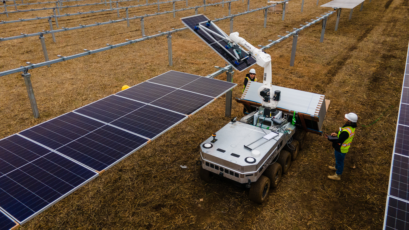 A mobile robotic system equipped with a KUKA robot positions solar panels on racking systems in an open field.